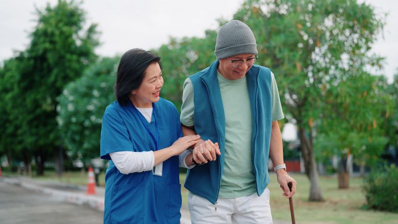 Older man with a walking cane is being supported by a female medical professional during a walk.