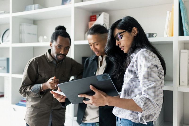 A diverse group of colleagues reviews documents on a tablet in a bright, shelves-filled workspace. They discuss ideas, share notes, and collaborate to solve a team task.