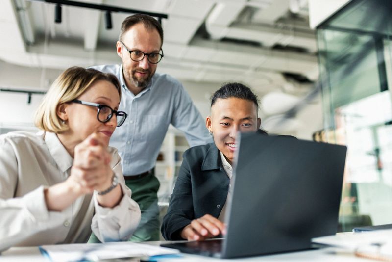 A diverse group of colleagues reviews data on a laptop in a bright, open-plan office. One person points at the screen while others listen, suggesting teamwork, planning, and discussion.