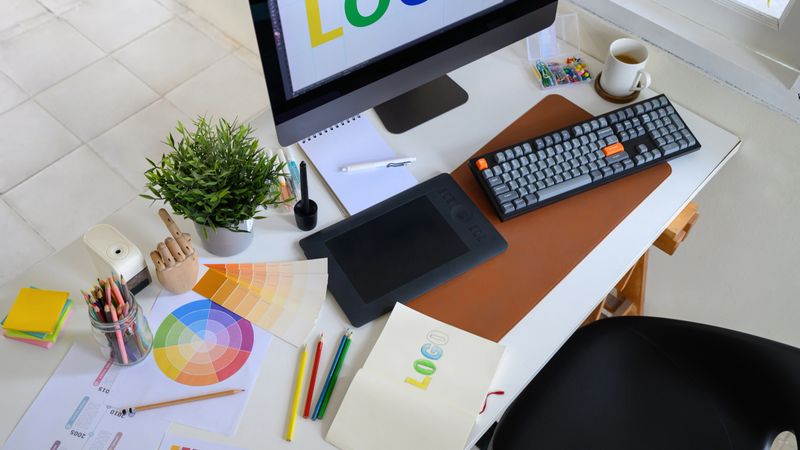 Creative woman designing logo sketches on notebook with color samples and tools in bright modern workspace.