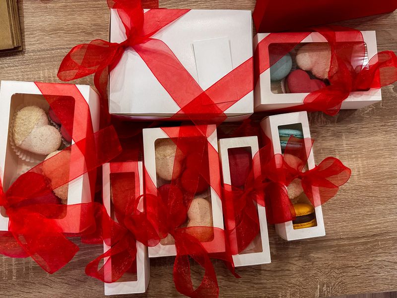 A charming assortment of individually packaged macarons and heart-shaped pastries, elegantly presented in white gift boxes adorned with vibrant red sheer ribbons, ready for a special occasion.
