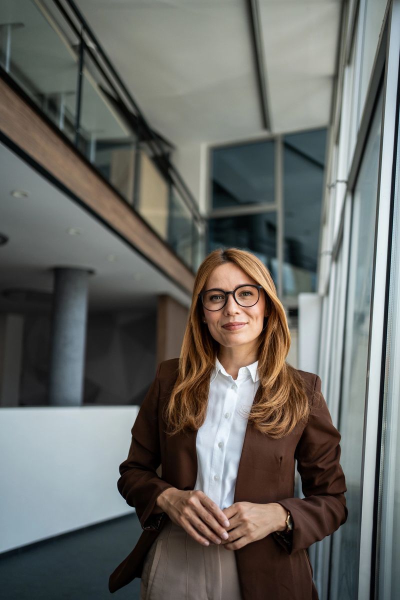 Businesswoman with glasses and brown jacket standing confidently in a bright, modern corporate office building, portraying professionalism, leadership, and success in a contemporary work environment