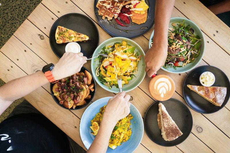 A lively overhead shot of a cozy brunch table adorned with waffles, salads, and coffee art, as hands reach in to enjoy the vibrant spread.