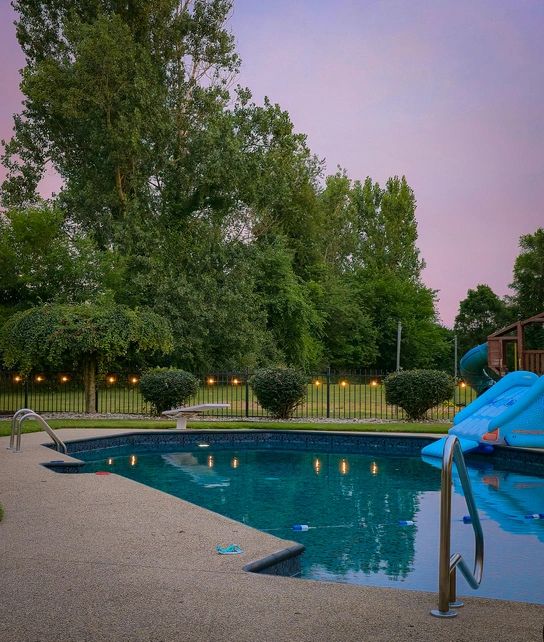 A serene backyard pool glows under string lights as dusk settles in. Surrounded by lush trees and a quiet play area, the calm water and peaceful ambiance create a relaxing summer evening scene.