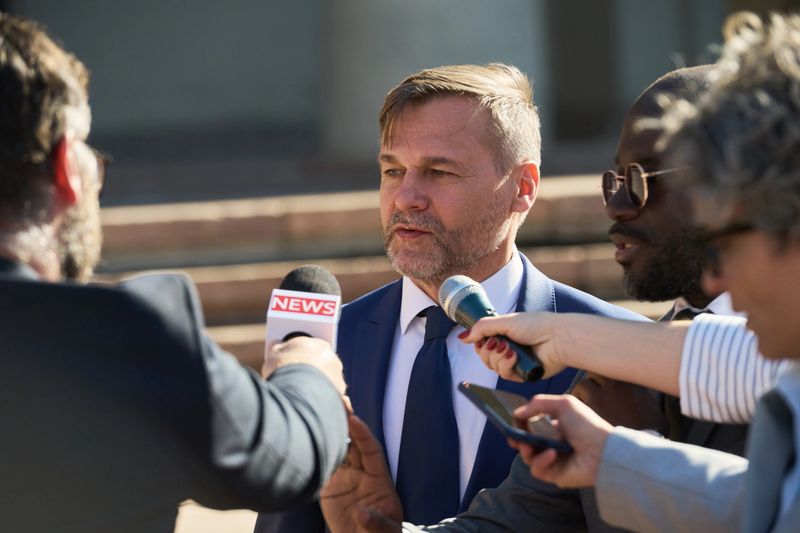 Caucasian middle aged man in suit speaking to group of journalists holding microphones and recording devices outdoors, Black man and other reporters surrounding subject during interview