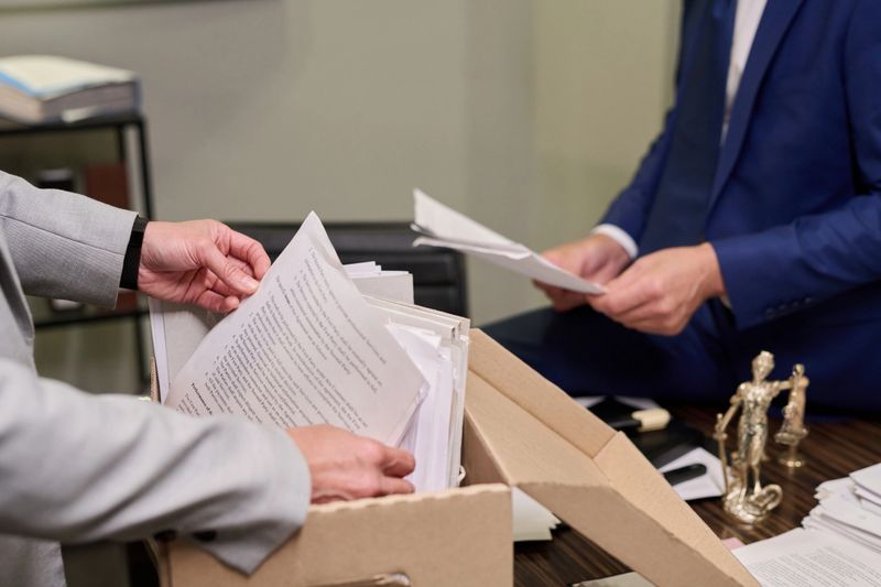 Middle aged Caucasian man in suit sorting legal documents at desk while another middle aged Caucasian man organizing paperwork from cardboard box, law office setting visible