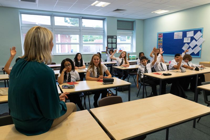 A wide over-the-shoulder shot of a smiling female teacher standing at the front of a bright, modern classroom, engaging a group of students. The students, dressed in matching white shirts and striped black ties, sit at their desks listening attentively while some raise their hands. The atmosphere is positive and interactive, highlighting effective teaching, learning, and communication in an academic environment.Videos are available similar to this scenario.