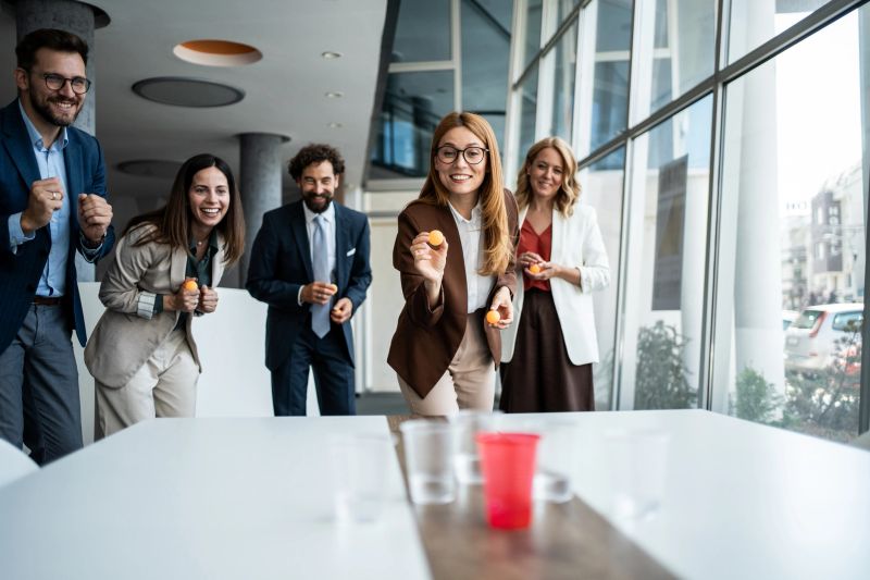Happy business colleagues are laughing and playing a friendly game of beer pong together during an office team building event, fostering good work-life balance and fun