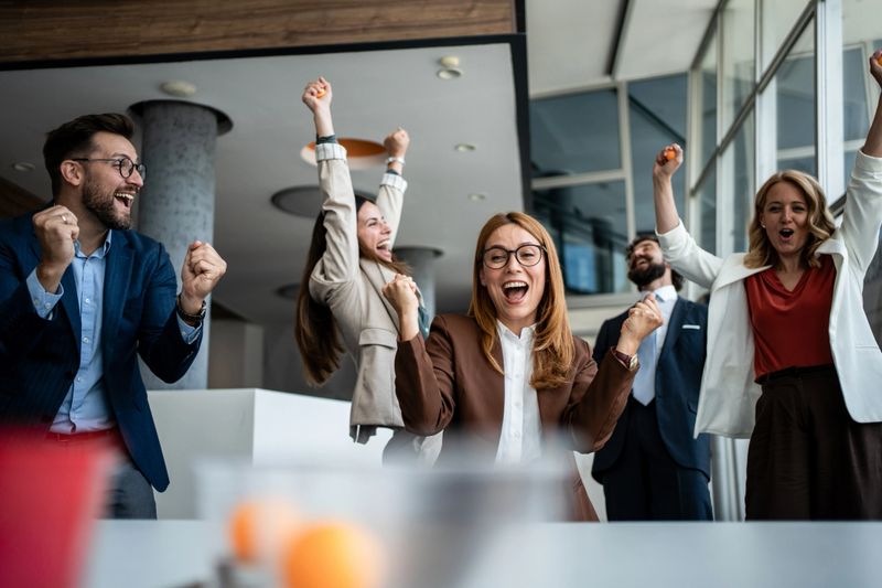 Group of happy business people celebrating victory and achievement together, raising their fists and smiling while standing in a contemporary office setting after a successful project