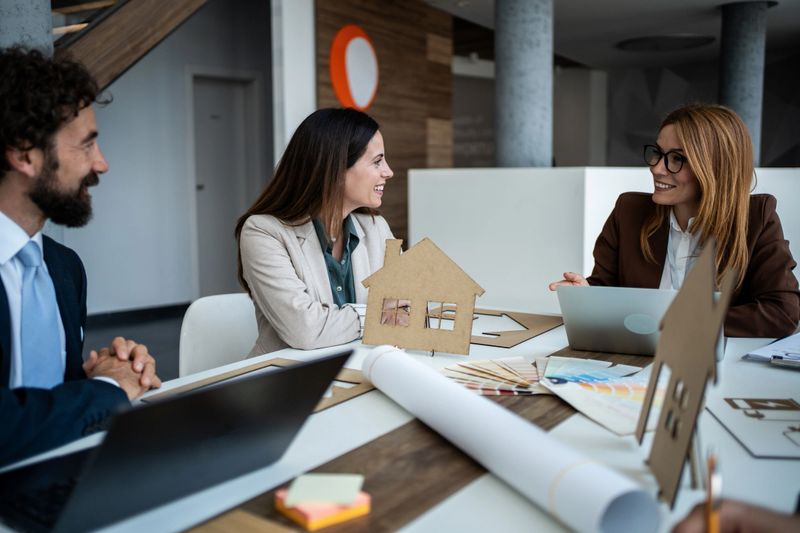 Architects and planners collaborating in an office around a scale house model, reviewing blueprints and discussing real estate design, development strategy, and project plans