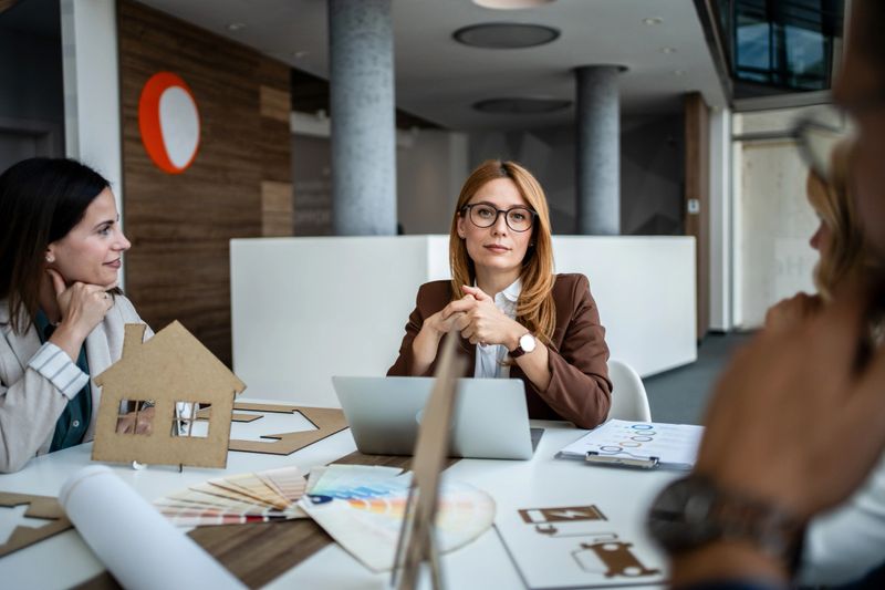 Businesswoman architect leading a meeting with colleagues, discussing house design and real estate project planning, using a cardboard house model and laptop on the office table