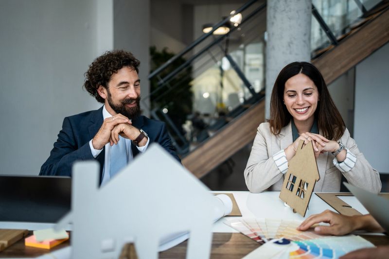 Two smiling architects discussing a new housing project, holding a house model and looking at color swatches, representing collaboration and planning in real estate