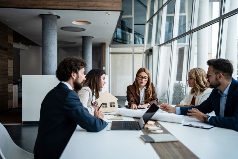 Group of diverse business professionals collaborating during a meeting in a modern office, reviewing blueprints and a house model while discussing real estate and architecture development plans