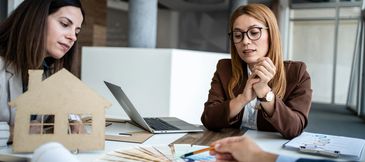 Two women discussing real estate plans at a desk with a laptop and house model.