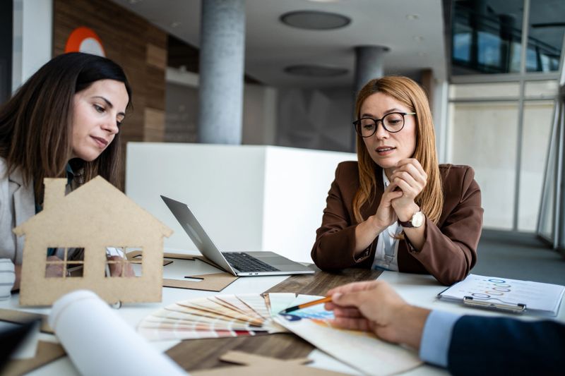 Female architects and a client are collaborating to select interior design elements, discussing a house model, color swatches, and building plans in a modern office