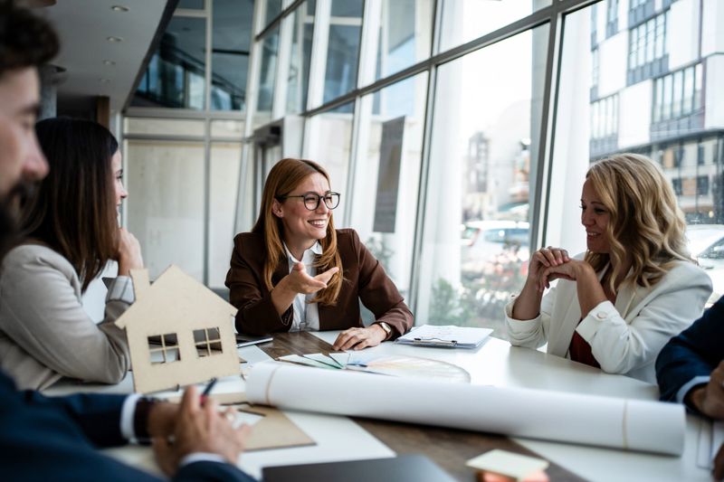 Diverse group of business people collaborating and discussing a real estate development project, with one woman presenting ideas while a house model sits on the table in a modern corporate office