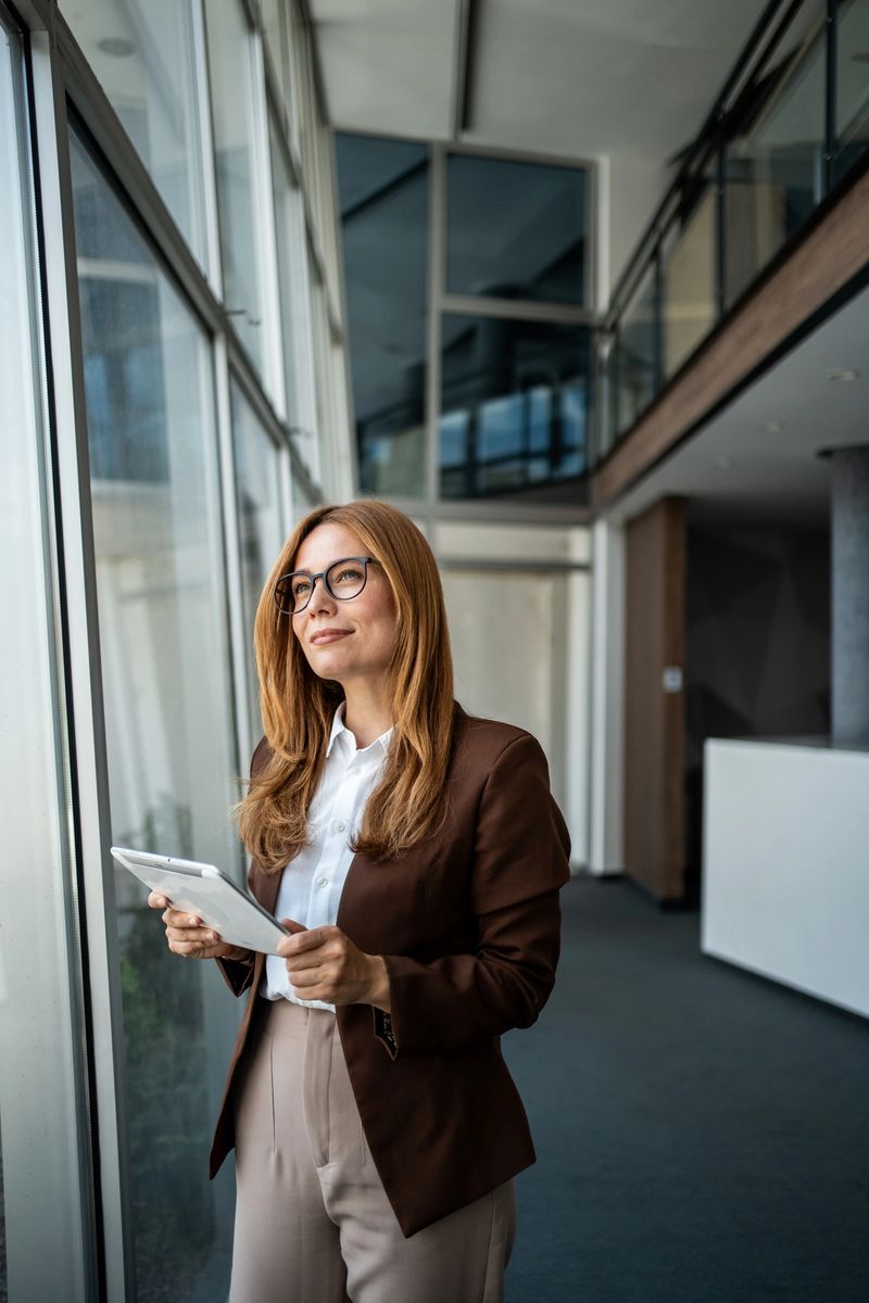 Professional businesswoman in corporate office looking through large windows and holding a digital tablet, reflecting on future vision and business success