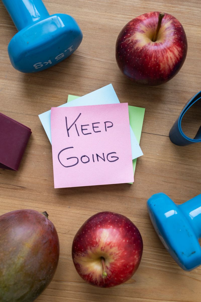 Green sticky note with handwritten Keep Going message placed on a table surrounded by fresh fruit and workout accessories, symbolizing motivation, perseverance, and a healthy active lifestyle.