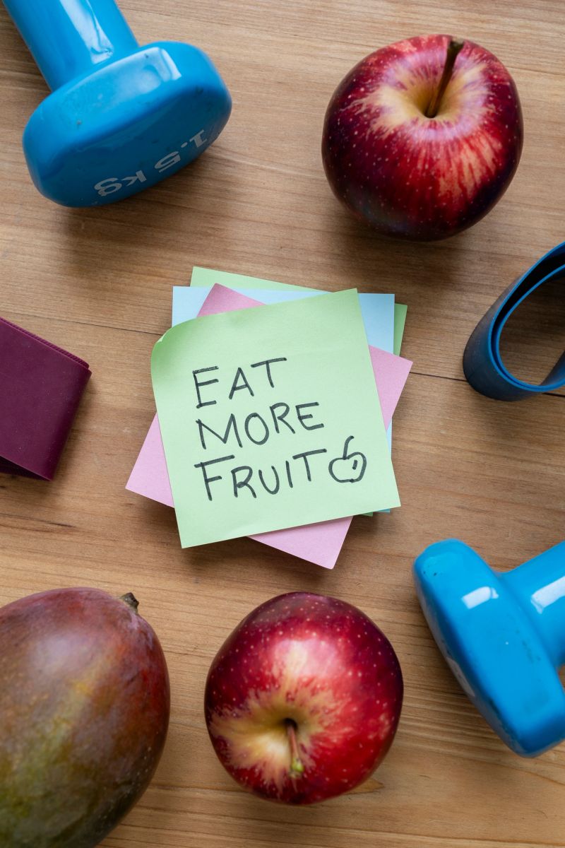 Green sticky note with handwritten Eat More Fruit message placed on a table surrounded by colorful fresh fruit and workout accessories, promoting healthy eating, wellness, and active lifestyle.