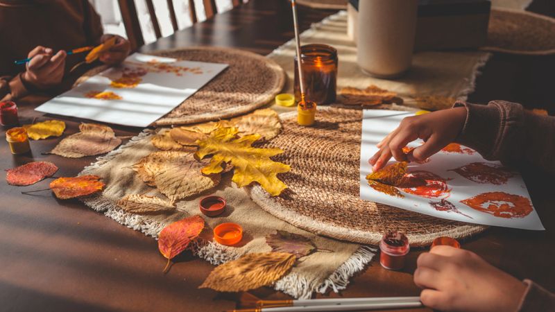 Children are engaged in an autumn-themed art project, using leaves as stamps with colorful paint on a table, creating a fun and educational activity.