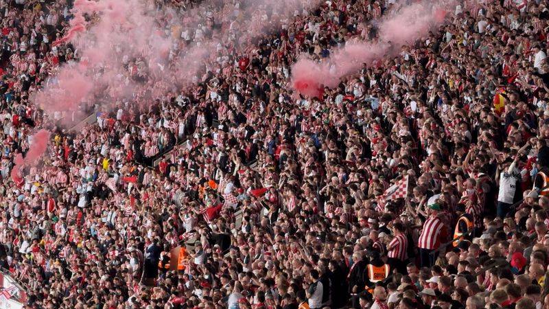 Energetic football fans fill the stadium podium, all dressed in red as they wave flags and ignite pink-red flares before the match begins. The atmosphere is electric — glowing smoke, roaring chants, and passionate unity create a stunning visual of team spirit. Captured in dynamic lighting and vivid colors, the scene reflects the intensity and emotion of European football culture.