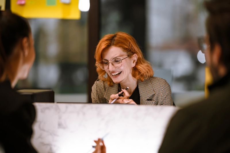 A woman with orange hair and glasses is happily interacting with two people at a stylish workspace. The environment is modern, with bright colors and a welcoming atmosphere.