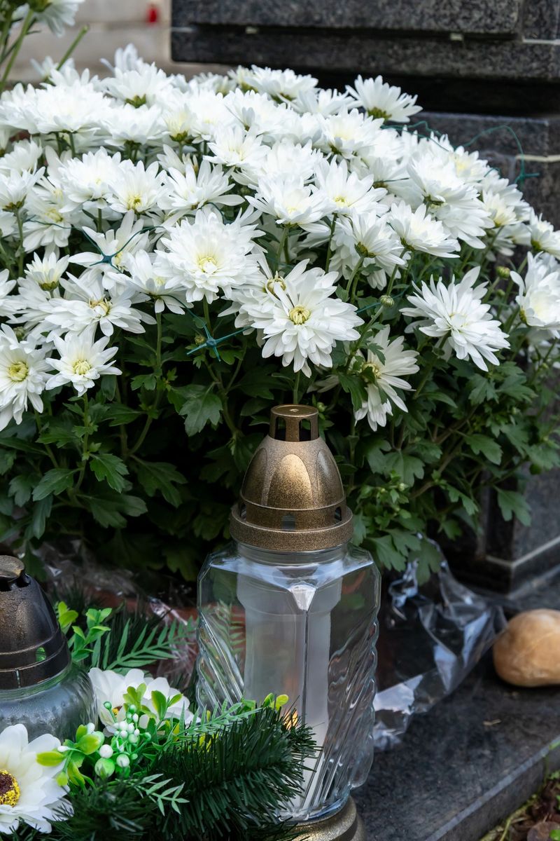 Close-up view of fresh white chrysanthemums placed beside glass cemetery candles on a gravestone. The soft candlelight and delicate flowers create a peaceful and respectful atmosphere, symbolizing love, remembrance, and eternal memory.