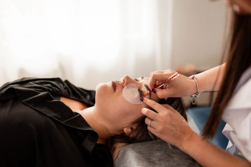 Cosmetologist applying dye and performing lash lift and eyebrow shaping for client in salon.