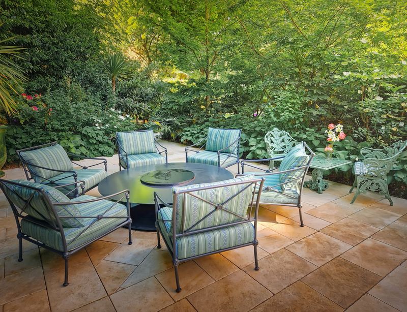 Elegant outdoor patio setting with circular table surrounded by cozy chairs with cushions. The space is framed by lush green foliage, creating a private, secluded, and luxurious garden atmosphere