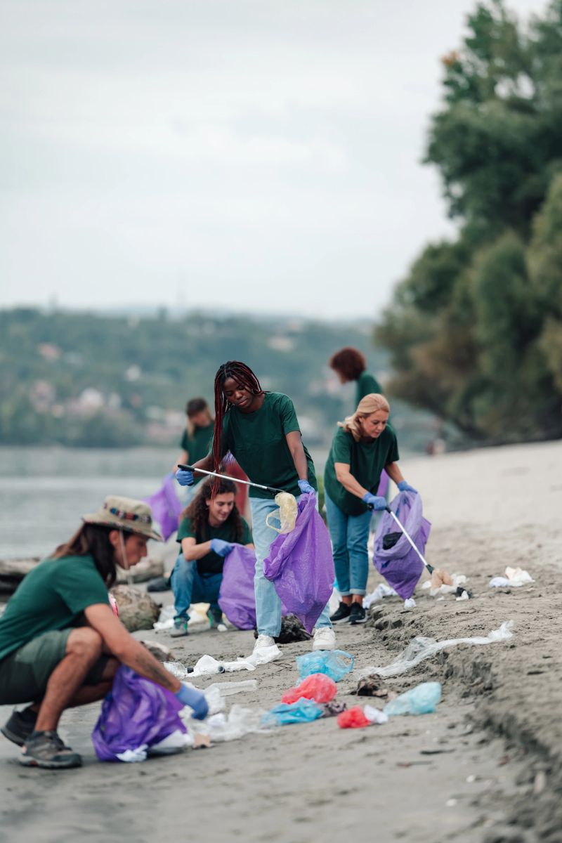 Group of volunteers wearing green t-shirts and gloves, actively picking up plastic waste and trash into purple bags on a sandy riverbank