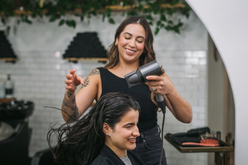 A smiling female stylist with tattoos blowing out a woman's hair with a hairdryer in a modern boutique salon.