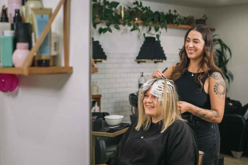 A smiling female stylist with tattoos folding hair foils for highlights on a smiling blond woman in a modern boutique salon.