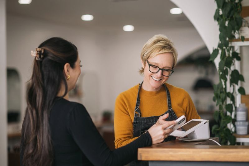 A happy female salon owner wearing an apron, ringing up a female client with tap-to-pay at the front desk of a modern boutique salon.