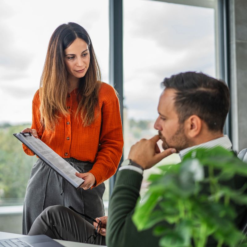 Young businesswoman explains project details to male colleague while holding clipboard with documents, collaborating in a modern office during an informal meeting and review