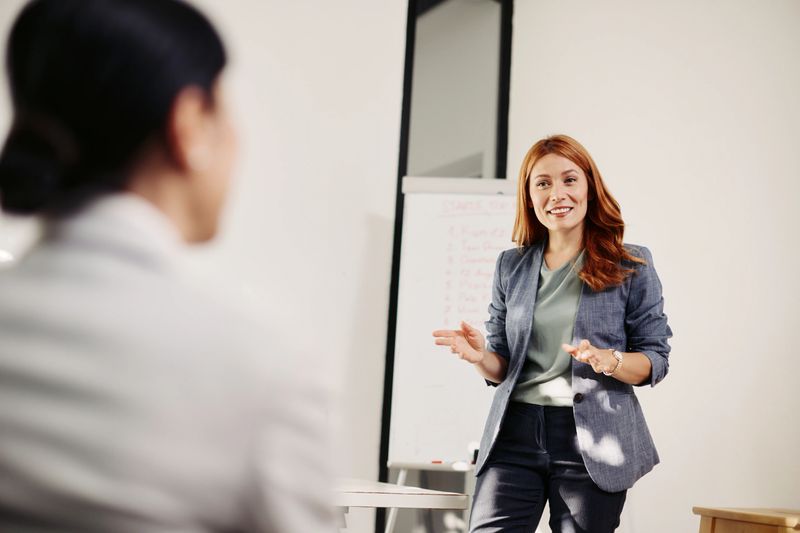 A confident speaker leads a seminar in a bright, modern conference room. A female presenter stands at the front with a flipchart, addressing a small group of professionals who are seated and listening intently. A laptop and flipchart are visible, suggesting a structured training or life coaching session. Natural light and clean, contemporary decor create a professional yet inviting learning environment.