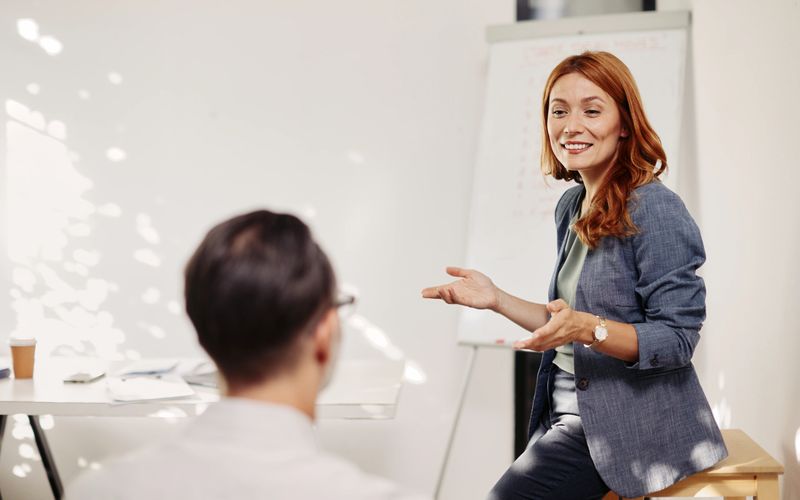 A confident speaker leads a seminar in a bright, modern conference room. A female presenter stands at the front with a flipchart, addressing a small group of professionals who are seated and listening intently. A laptop and flipchart are visible, suggesting a structured training or life coaching session. Natural light and clean, contemporary decor create a professional yet inviting learning environment.