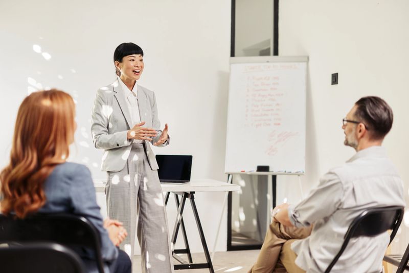 A poised female presenter of Japanese descent stands at the front of a bright, minimalist room, delivering a training or workshop. She uses expressive hand gestures while two participants sit in front, listening attentively. A laptop, notebooks, and a flip chart are visible, suggesting an interactive learning moment. Natural light and clean, modern surroundings create a professional and inviting environment. Model release is on file.