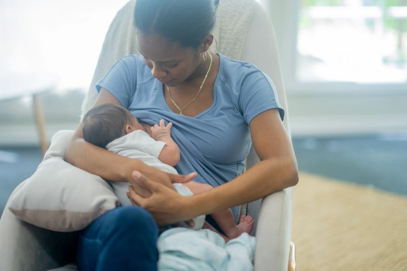 A loving Black mother sits in a soft chair, nursing her newborn close. The intimate moment highlights maternal bonding, warmth, and family care in a bright, tranquil home.