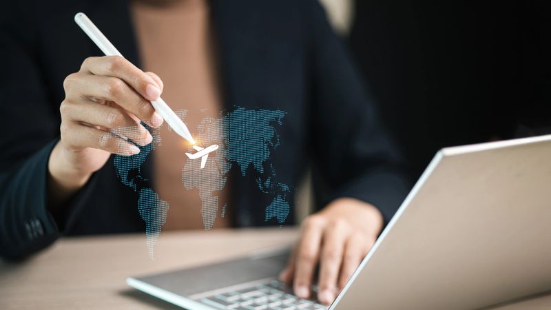 Businessperson using stylus to select airplane icon on virtual world map above laptop. Concept for global travel, logistics technology, online booking, smart tourism, and business connectivity.