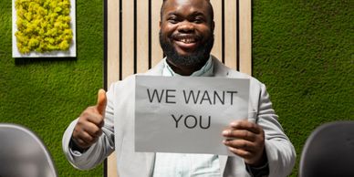 Smiling man holding a 'WE WANT YOU' sign and giving a thumbs up.