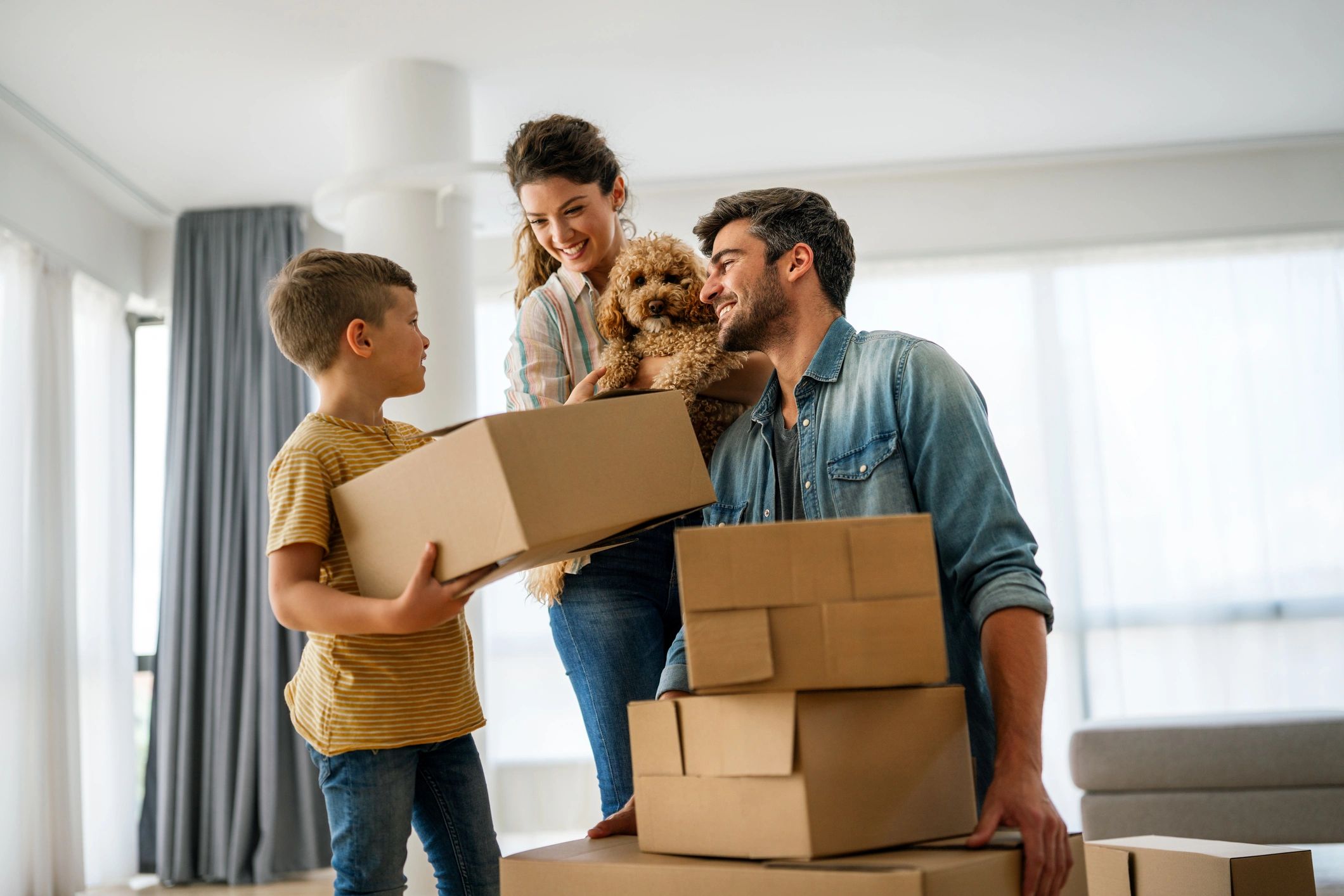 A smiling family of three moving into a bright new home with their small brown dog. The young boy ho