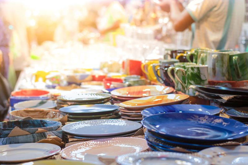 The image was captured on September 25, 2025, inside a busy market shop in Gaibandha, Bangladesh. The scene shows a male seller actively engaging with buyers, most notably women in traditional attire. The shop is overflowing with a vast and colorful array of kitchen accessories and household goods, including metal pots, silverware, glass cups, and stacks of vibrant plates in the foreground. This bustling environment vividly illustrates the energy of local commerce, with the seller working to serve his community by providing essential kitchenware and decorative items.