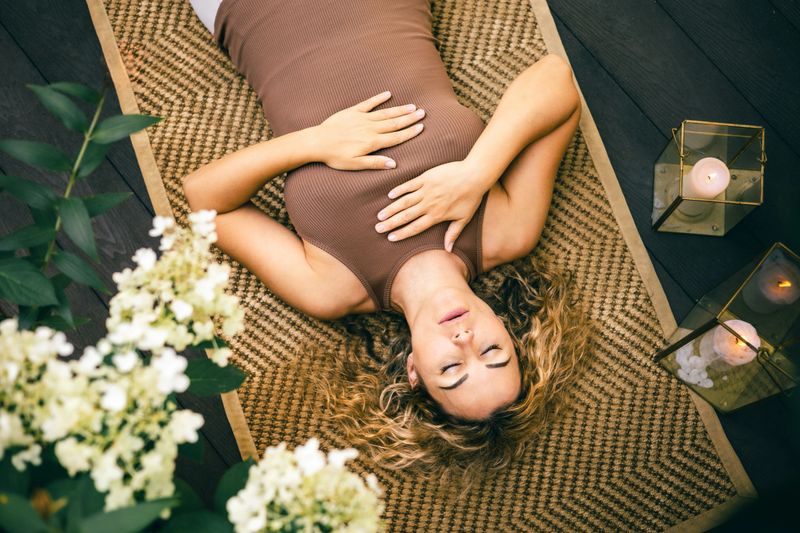A woman lies on a woven yoga mat outdoors with hands on her chest in shavasana, eyes closed, relaxing near candles and flowers in a calm peaceful atmosphere