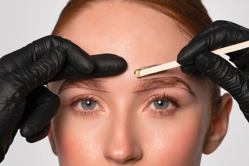 A skilled beauty professional is applying warm wax on a client's eyebrows in a clean and bright salon. The focus is on precise shaping and attention to detail.