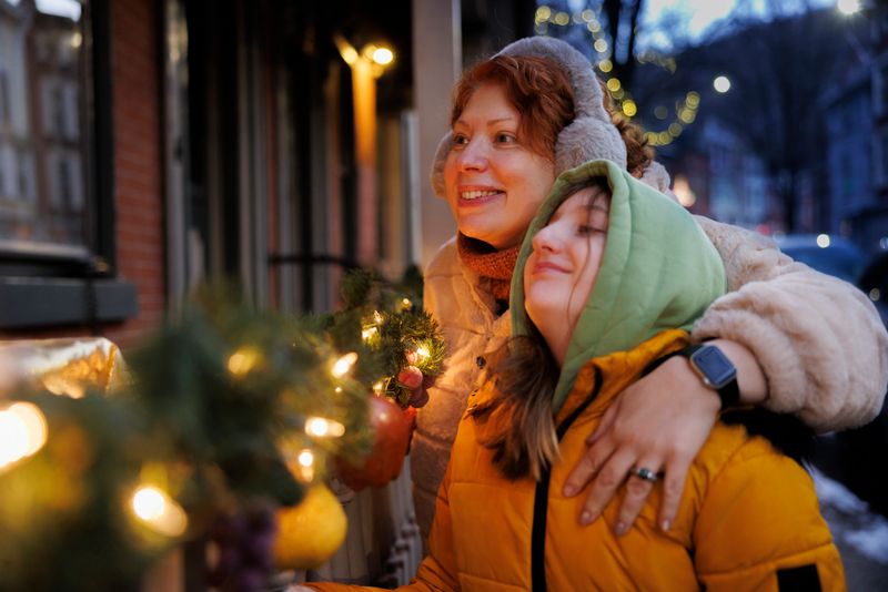 A redheaded middle-aged woman embraces her preteen daughter as they admire Christmas decorations outside a storefront in Jim Thorpe, Pennsylvania. The mother smiles warmly while her daughter leans affectionately against her, both illuminated by the soft glow of holiday lights. The evening scene captures their close bond during a winter outing in the historic Poconos town. Decorative evergreen branches with twinkling lights create a festive foreground, with the charming brick buildings of the downtown area visible in the background. The close-up shot focuses on their expressions of joy and contentment during this seasonal family moment.