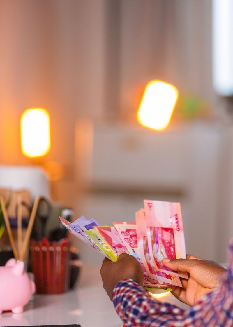 A person counts colorful Ghanaian cedi banknotes at a desk under warm indoor lighting, with a blurred background of lamps, stationery, and a pink piggy bank suggesting savings or finance.