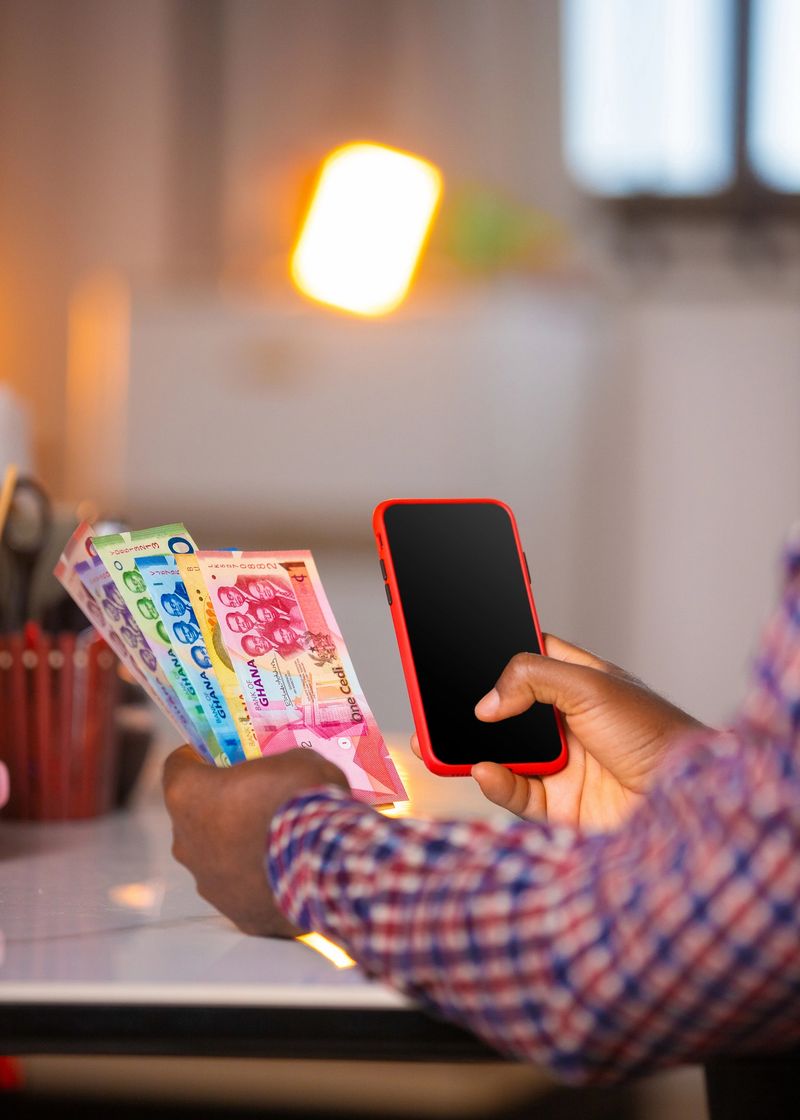 Black African man holding  colorful Ghanaian cedi banknotes and smartphone at a desk under warm indoor lighting, with a blurred background of lamps,  and stationery suggesting savings or finance
