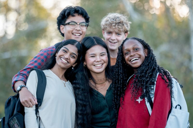 A cheerful, diverse group of five friends stands close together outdoors, smiling at the camera. They wear casual clothes and backpacks, suggesting campus life, friendship, and positive vibes among peers.