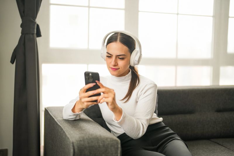 Young woman wearing wireless headphones using smartphone while relaxing at home — listening to music or podcast online.