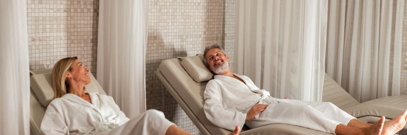 A middle-aged couple relaxes in a spa lounge wearing white robes.